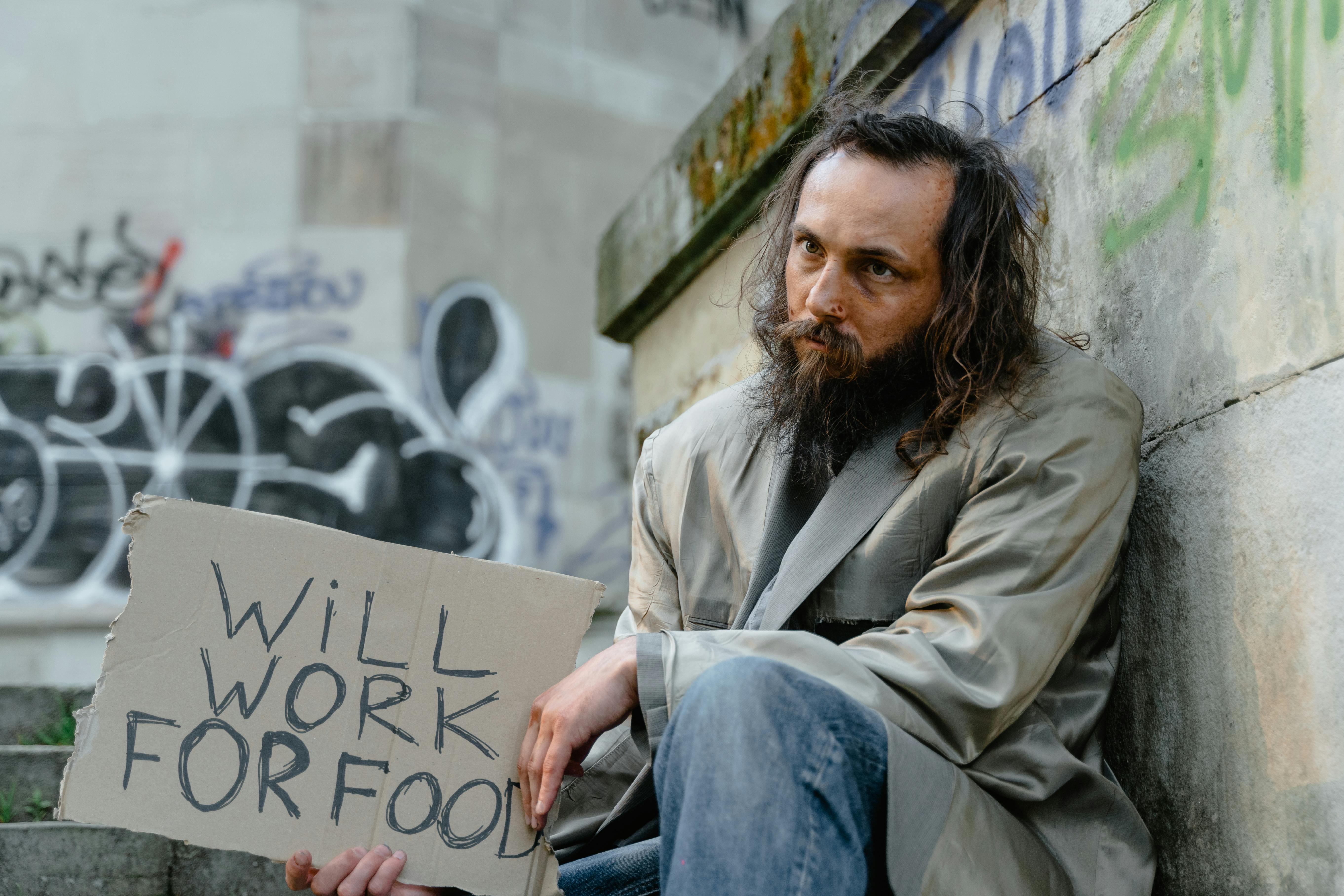 A homeless man with a sign expressing his willingness to work for food, sitting in an urban setting.