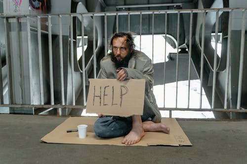 A Male Beggar Sitting on a Ground while Holding a Help Banner