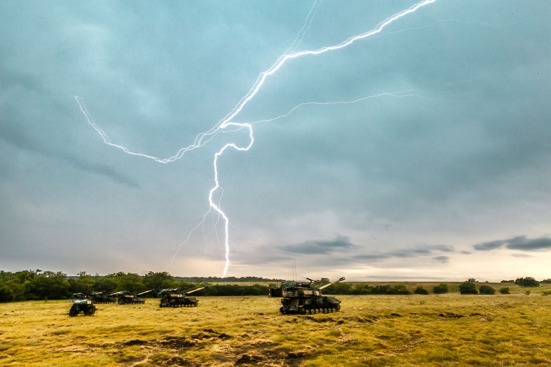 Dramatic lightning bolt striking above tanks in an open field under cloudy skies.