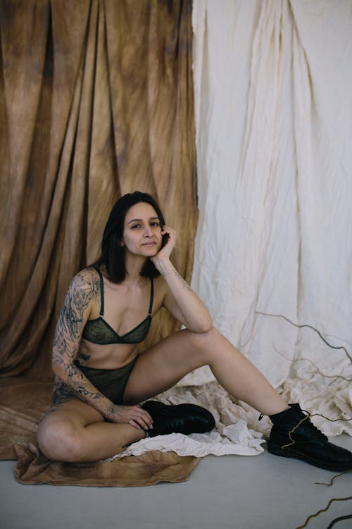 Full body of young seductive tattooed female wearing black lingerie and black leather boots sitting on floor on background of draped curtains
