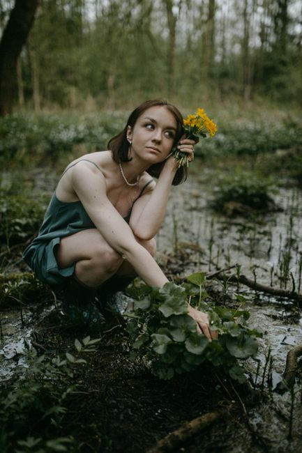 A woman wearing a green dress and rubber boots, holding yellow flowers, crouches in a swampy forest area.
