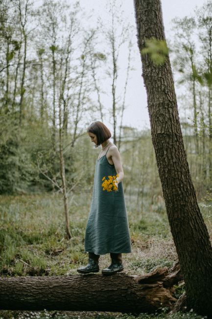 Woman in green dress holding yellow flowers while standing on a fallen tree in a serene forest.
