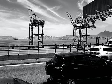 Black and white image of a bustling harbor with cranes, cars, and cityscape.