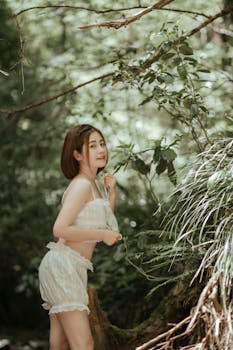 A young woman poses gracefully in a white outfit surrounded by lush forest greenery.