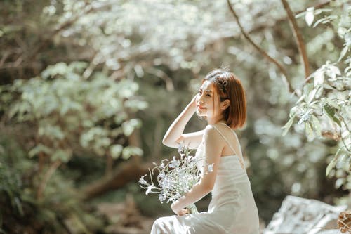 Free Pretty Woman in White Spaghetti Strap Dress Holding Fresh Flowers while Posing Stock Photo
