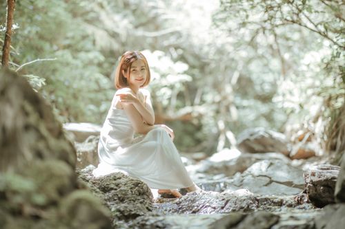 Free A woman in a white dress sits on rocks by a forest stream, enjoying natural tranquility. Stock Photo