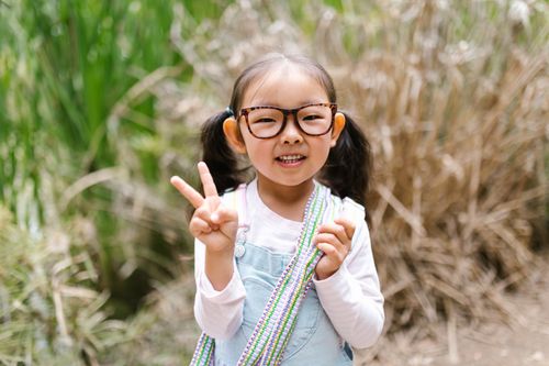 Free Cheerful Asian girl wearing glasses, making a peace sign in an outdoor setting. Stock Photo