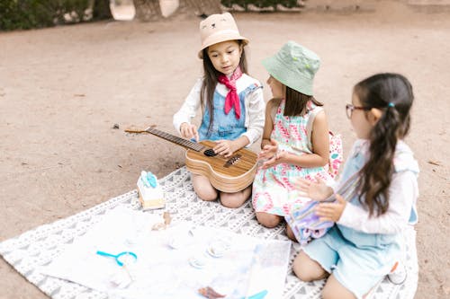 Free Girl in Denim Jumper with a Ukelele Sitting on Picnic Blanket Beside Two Girls Stock Photo