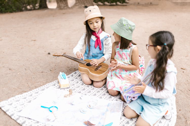 Girl In Denim Jumper With A Ukelele Sitting On Picnic Blanket Beside Two Girls