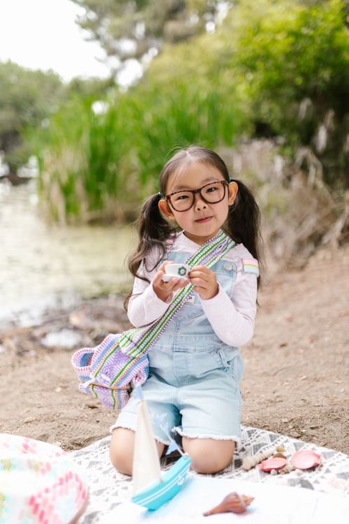 Free A young girl enjoys outdoor playtime with a toy camera by a lake, surrounded by nature. Stock Photo