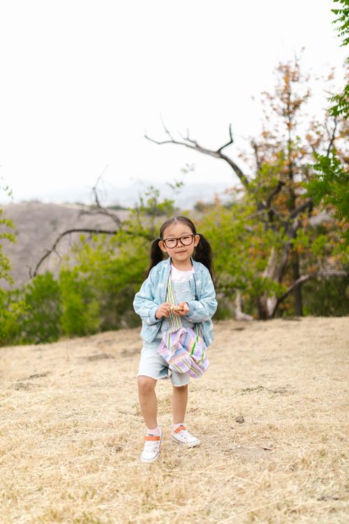 Free A young girl with glasses standing in a field holding a colorful backpack, surrounded by nature. Stock Photo