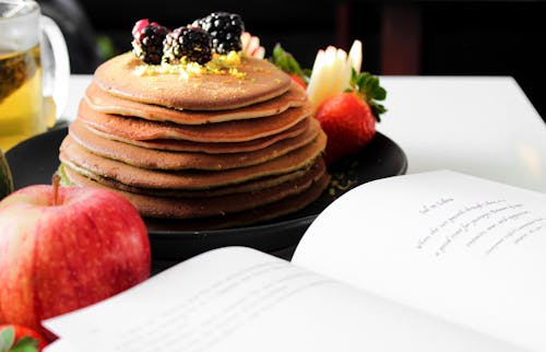 Delicious appetizing pancakes on black plate with blackberries on top and apple near opened book