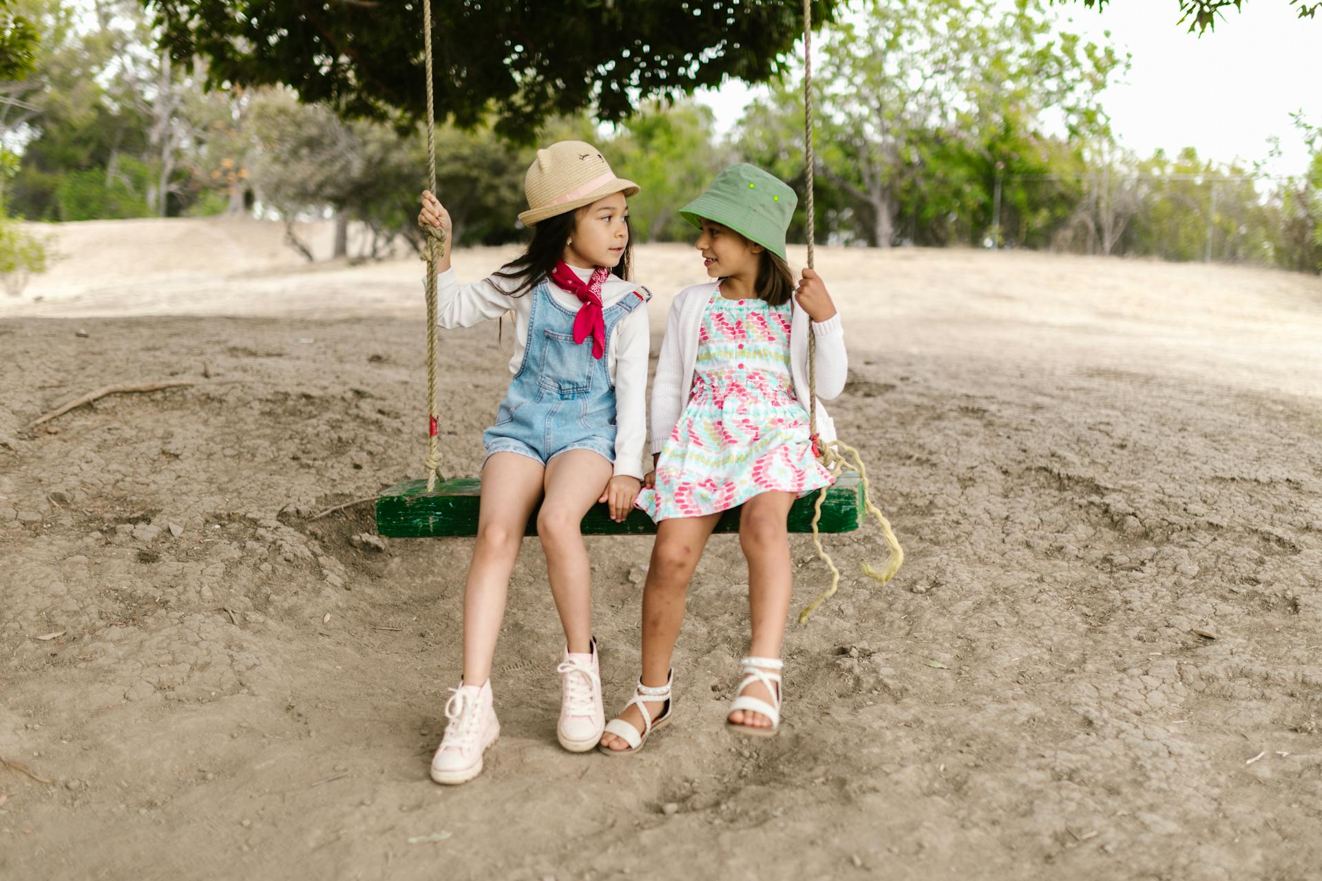 Two young girls in bucket hats share a joyful swing ride in a lush outdoor setting.