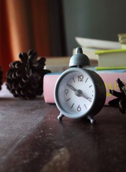 A small gray alarm clock on a wooden table surrounded by pine cones.