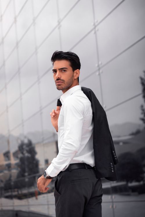 Free Confident young adult in white shirt and suit jacket posing outdoors against a reflective wall. Stock Photo