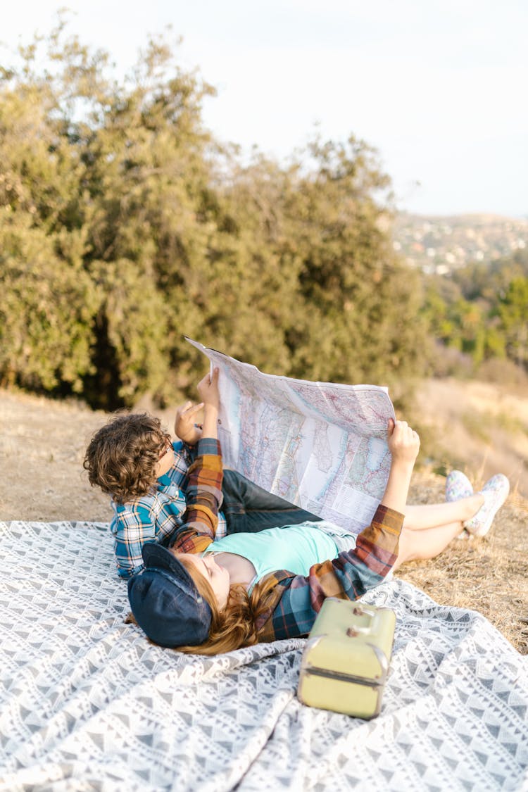 Two Kids Lying Down On Picnic Blanket While Looking At A Map