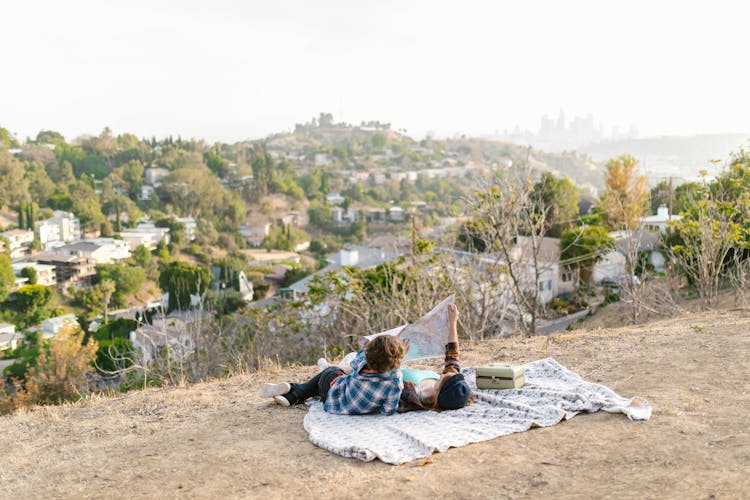 Two Teens Lying Down On Picnic Blanket While Looking At A Map