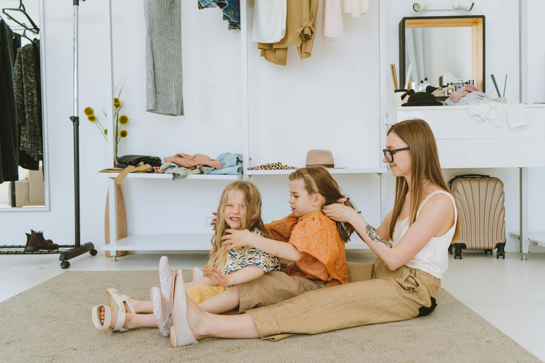 A loving mother braiding her daughters' hair in a bright, cozy room full of clothing and warmth.