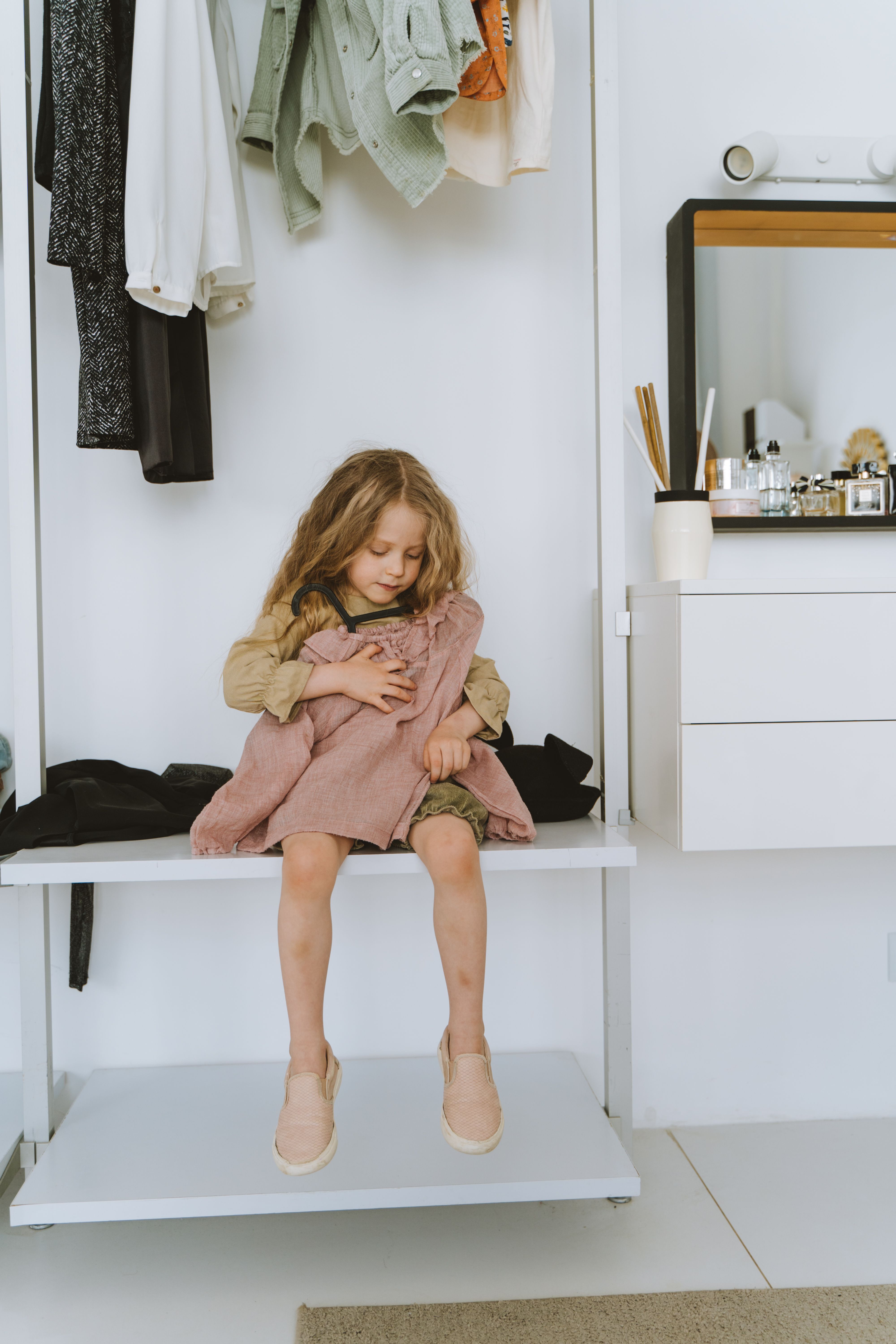 A young girl sitting and holding a pink blouse in a modern, minimalist closet setting.