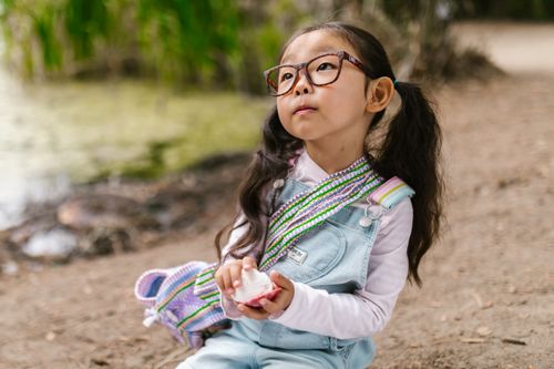 Free Cute young girl with pigtails and glasses holding a sea shell near a pond wearing a denim jumper. Stock Photo