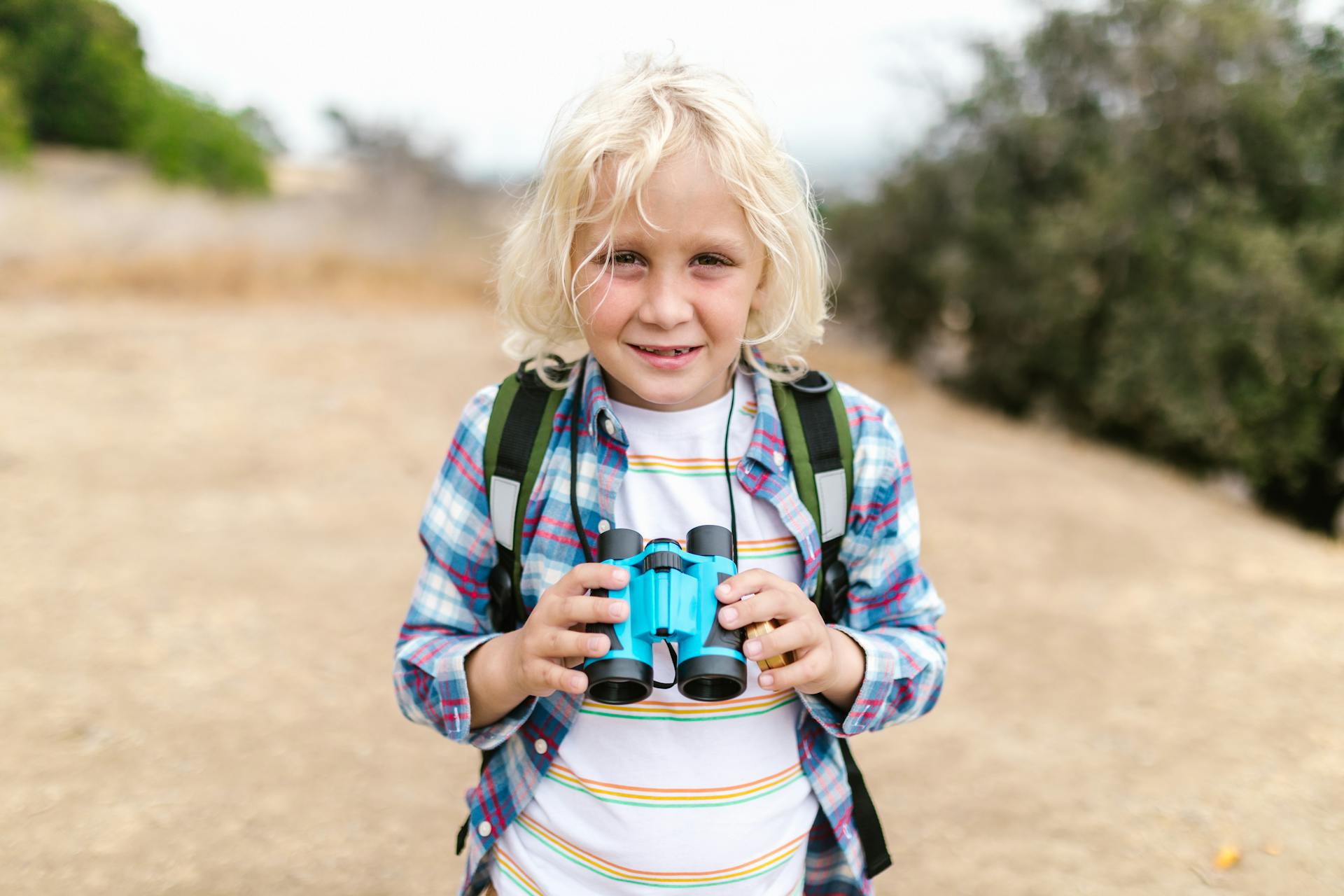 A young blonde child in a plaid shirt holds binoculars while exploring outdoors.