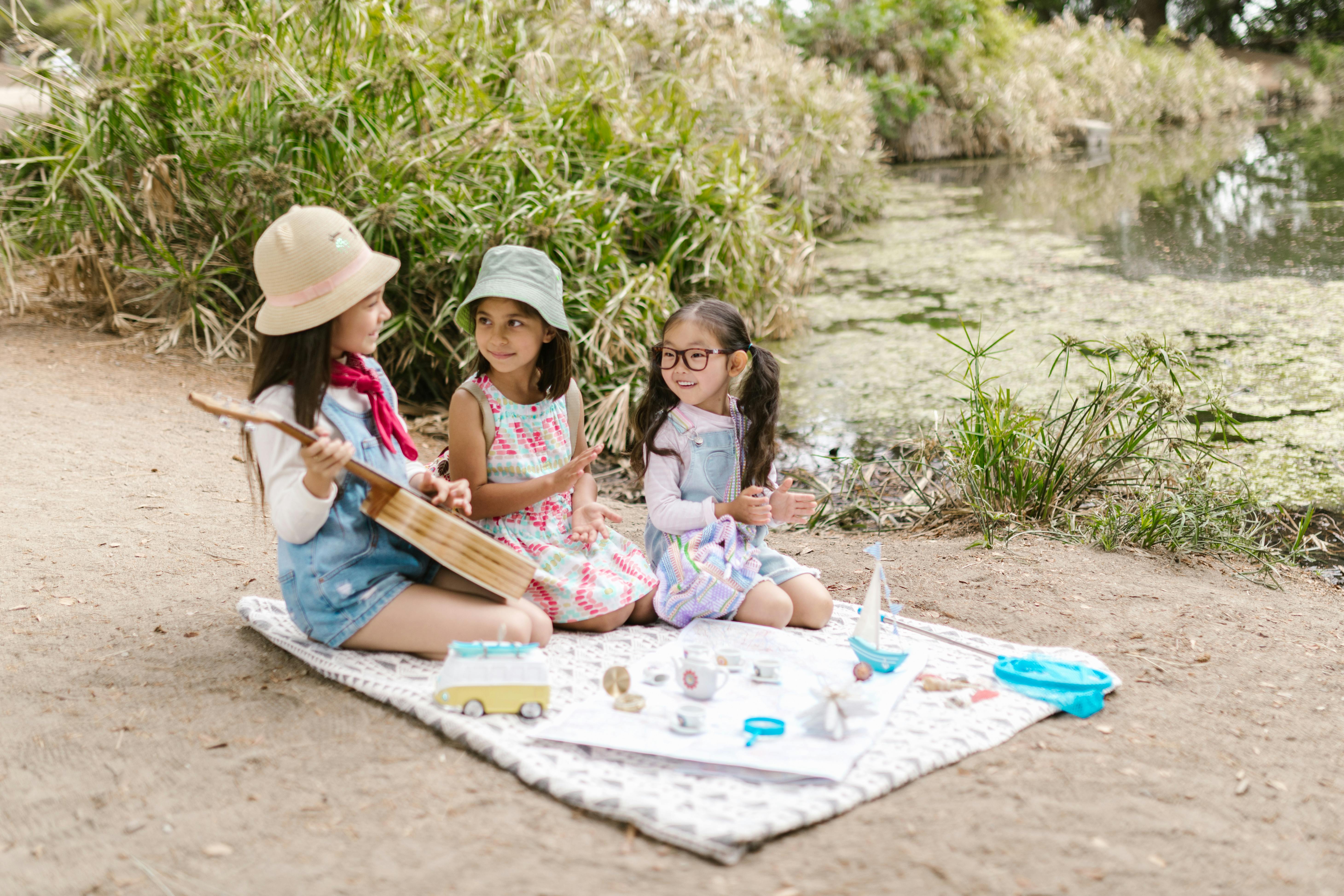 Premium Photo  Portrait of glad three kids sitting on blanket in field  playing ukulele having picnic with drinks and tasty food