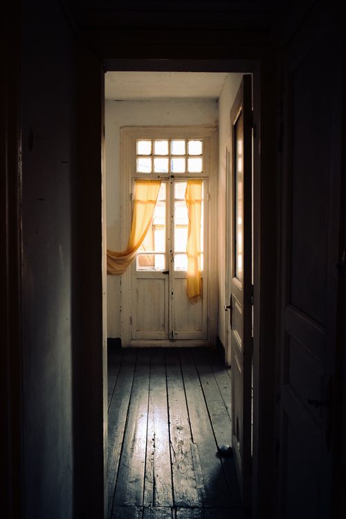 Free A dark, narrow hallway with wooden floors and sunlight filtering through yellow curtains on a door. Stock Photo