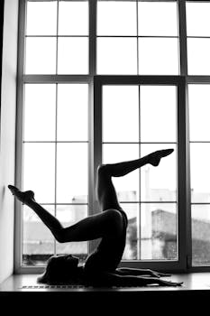 Black and white silhouette of a ballerina gracefully posed by a window, showcasing elegance and flexibility.