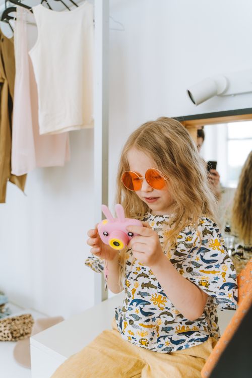 Free Blonde girl in sunglasses playing with a pink toy camera indoors, showcasing playful fashion. Stock Photo