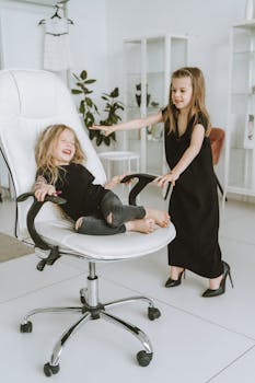 Two young girls playfully spinning on an office chair indoors, having fun together.