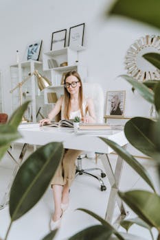 Woman reading a book at a desk in a modern office, surrounded by plants.