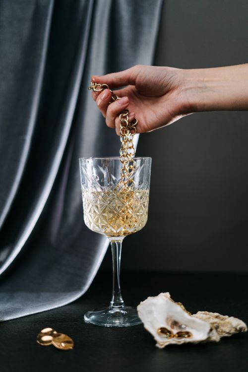 Free A woman's hand holding a gold necklace over a wine glass on a dark background. Stock Photo