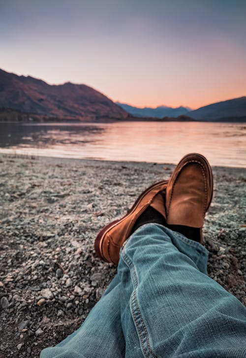 Person Wearing Blue Jeans and Brown Leather Loafers Sitting Beside Gray Sand