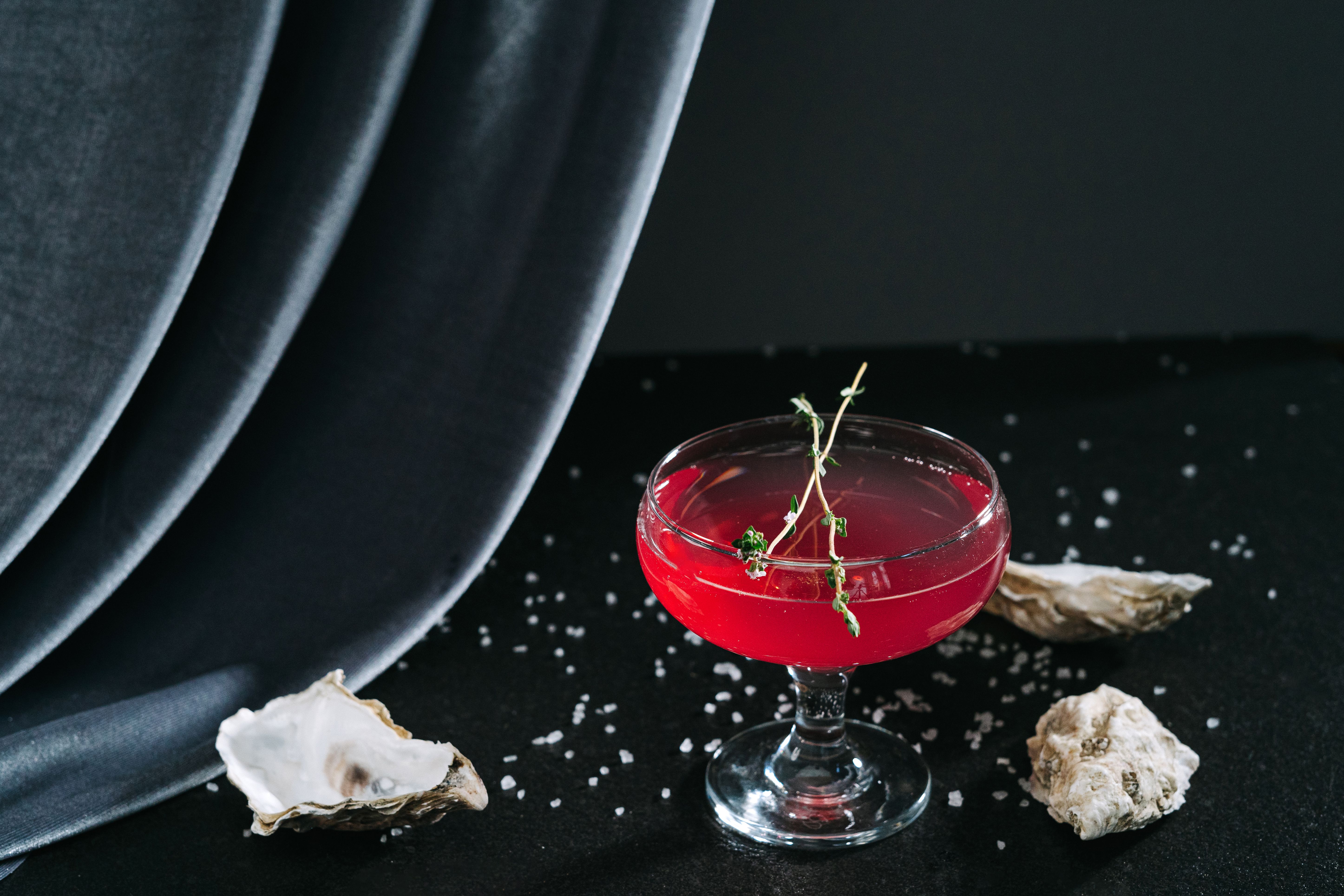 Deep oxblood-red cocktail in a coupe garnished with herbs on a glossy black surface, elegant and dramatic
