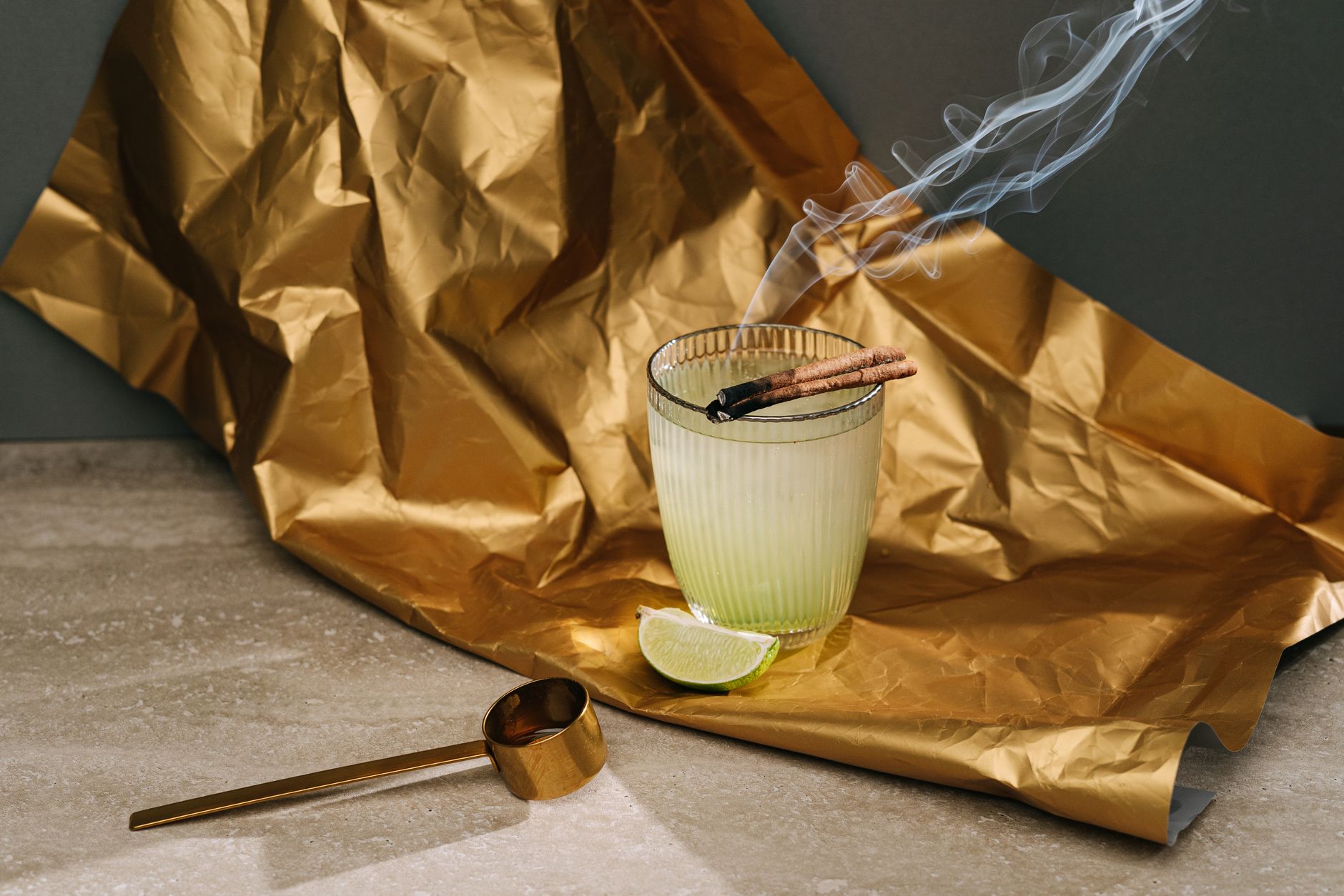 A cocktail with a cinnamon stick smoking over the glass, on gold foil