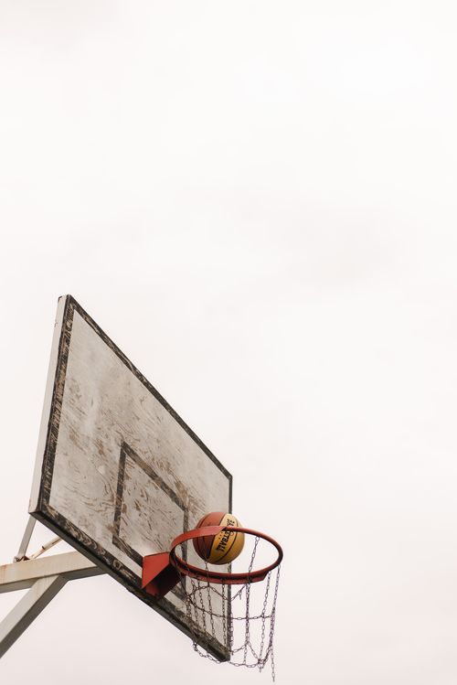 Free A basketball on the rim of a worn outdoor basketball hoop against a cloudy sky. Stock Photo