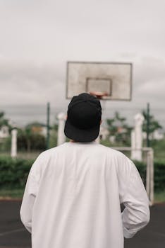 Person in white sweater and black cap looking at a basketball hoop outdoors.