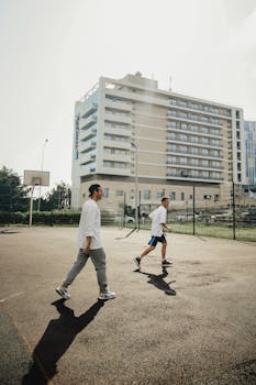 Two men play basketball on an outdoor court with a city building backdrop.