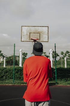 Rear view of a man in a red shirt aiming at a basketball hoop outdoors.