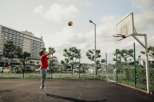 Man in red shirt playing basketball on an outdoor court, cityscape backdrop.