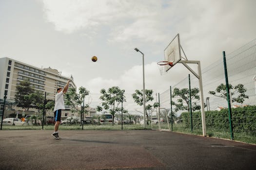 An adult male shooting a basketball on an outdoor court on a sunny day with buildings in the background.