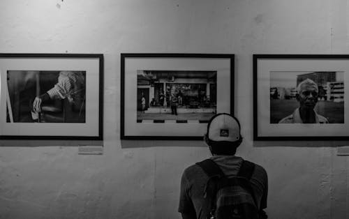Free Grayscale Photo of Man Wearing White Cap in Front of Three Paintings Stock Photo
