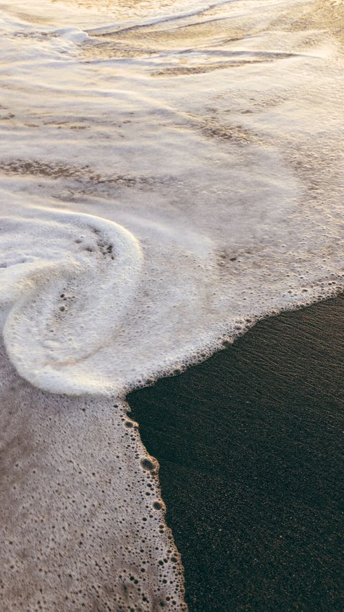 Free Foamy ocean waves gently crash on a sandy shore at sunset creating a serene landscape. Stock Photo