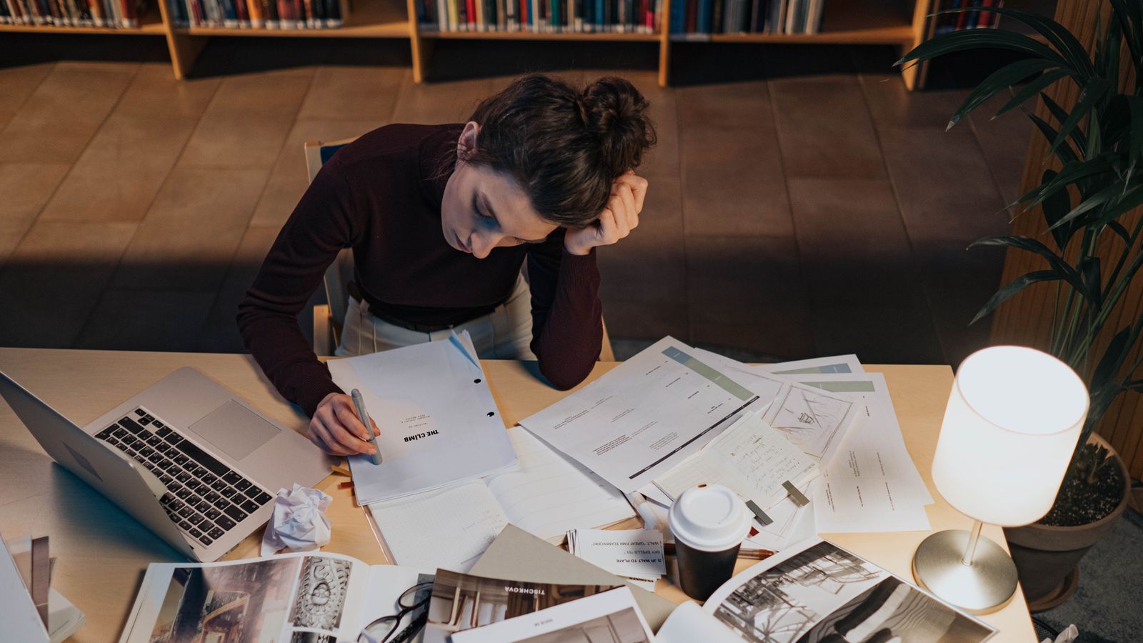 A woman reviewing printed pages at a desk with a laptop and coffee in a library setting