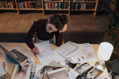 Free Woman studying at desk with documents, laptop, and coffee in a cozy library setting. Stock Photo