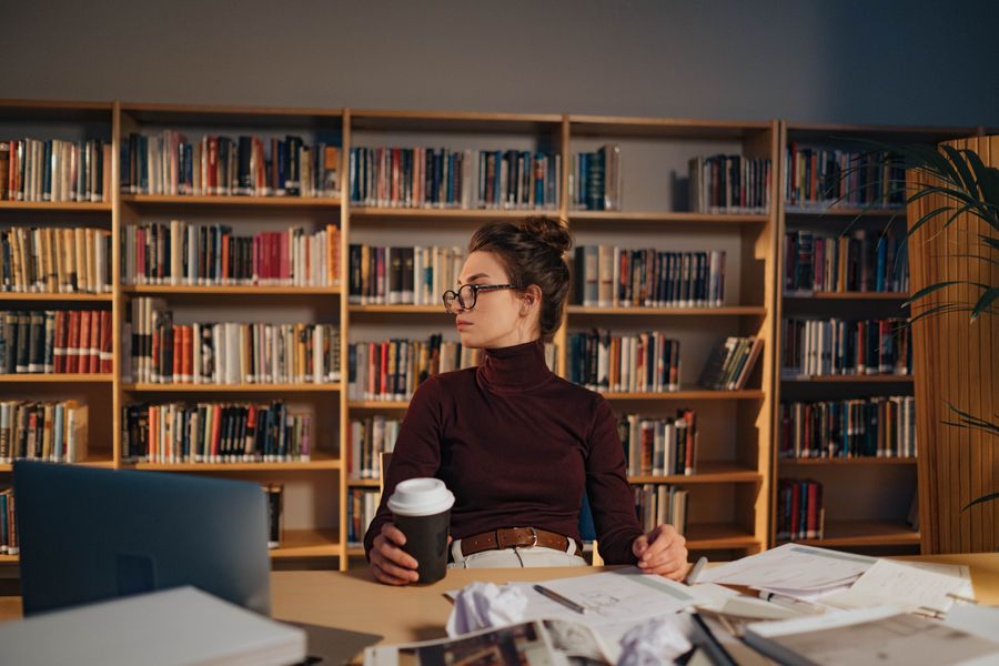 Brown-haired student with glasses studying with books and a laptop in a library