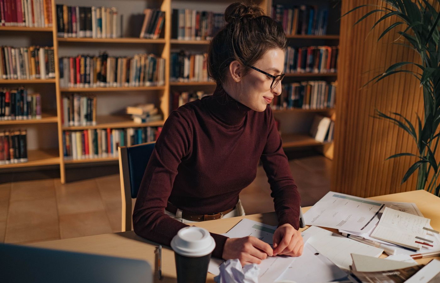 A woman in a library studying papers with determination, eyeglasses enhancing her focus.