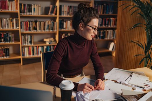 A woman in a library studying papers with determination, eyeglasses enhancing her focus.