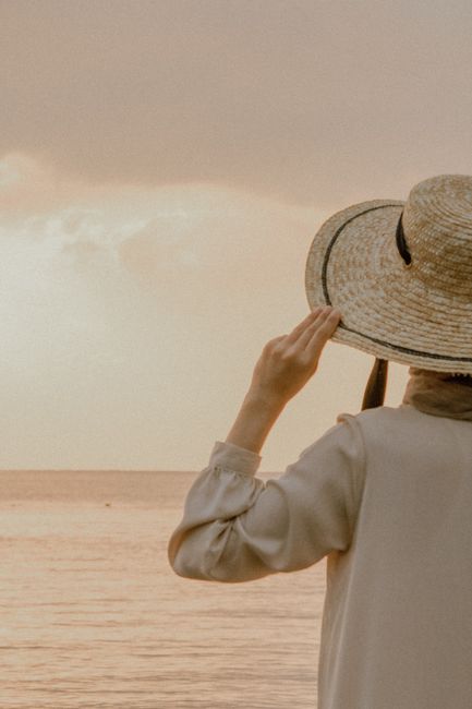 Woman in beige dress and straw hat admiring a tranquil seascape at sunset.