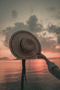 A hand holds a straw hat against a vibrant sunset reflected on the ocean waters.